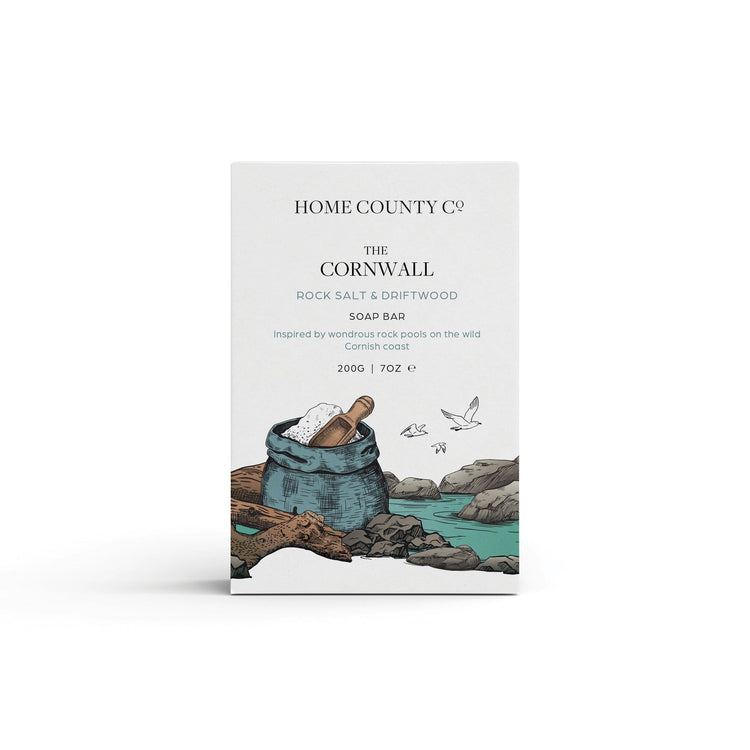 A rock salt and driftwood coastal scented natural soap bar from the Home County Co. is shown in its coastal illustrated recyclable soap bar wrap