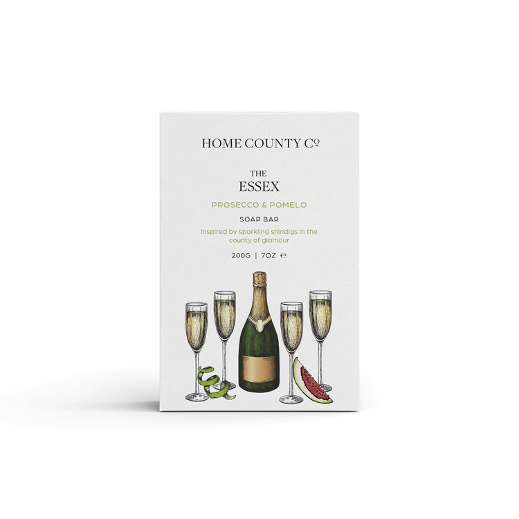 A prosecco scented natural soap bar from the Home County Co. is shown in its prosecco illustrated recyclable soap bar wrap