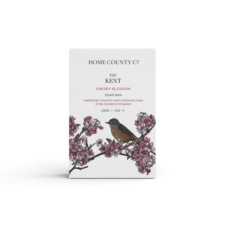 A cherry blossom floral scented natural soap bar from the Home County Co. is shown in its cherry blossom illustrated recyclable soap bar wrap