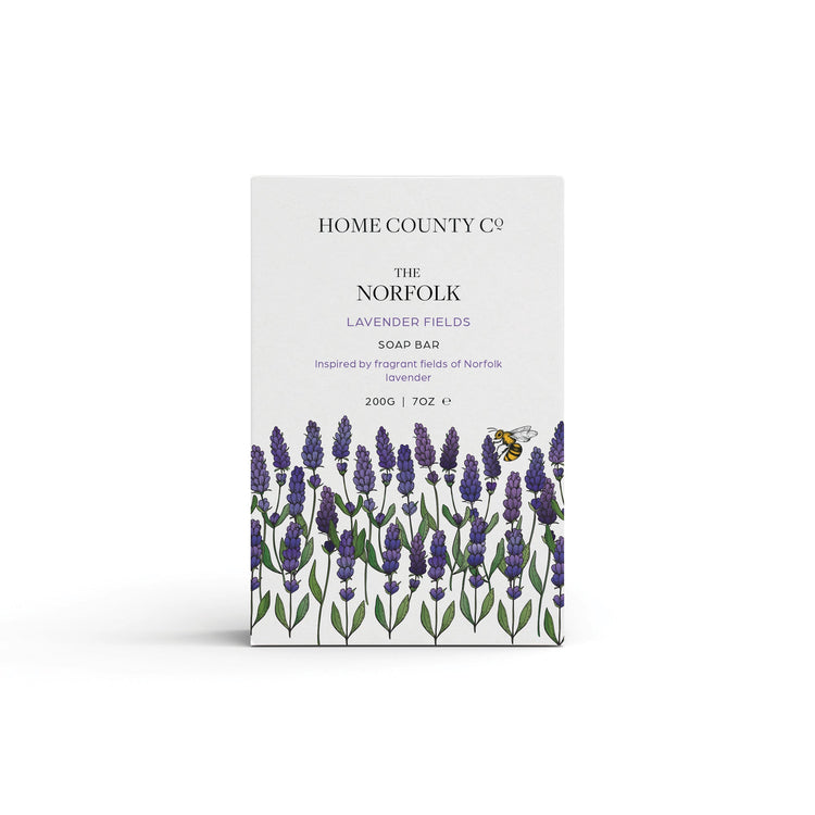 A Norfolk lavender scented natural soap bar from the Home County Co. is shown in its lavender illustrated recyclable soap bar wrap