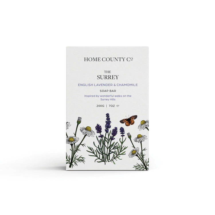 An english lavender and chamomile scented natural soap bar from the Home County Co. is shown in its lavender illustrated recyclable soap bar wrap