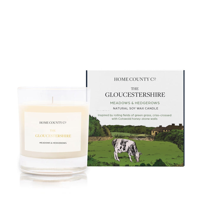 A freshly cut grass scented candle from Home County Co. The cotton wick soy candle is shown next to the eco friendly candle box packaging which displays an illustration of Cotswold hills.