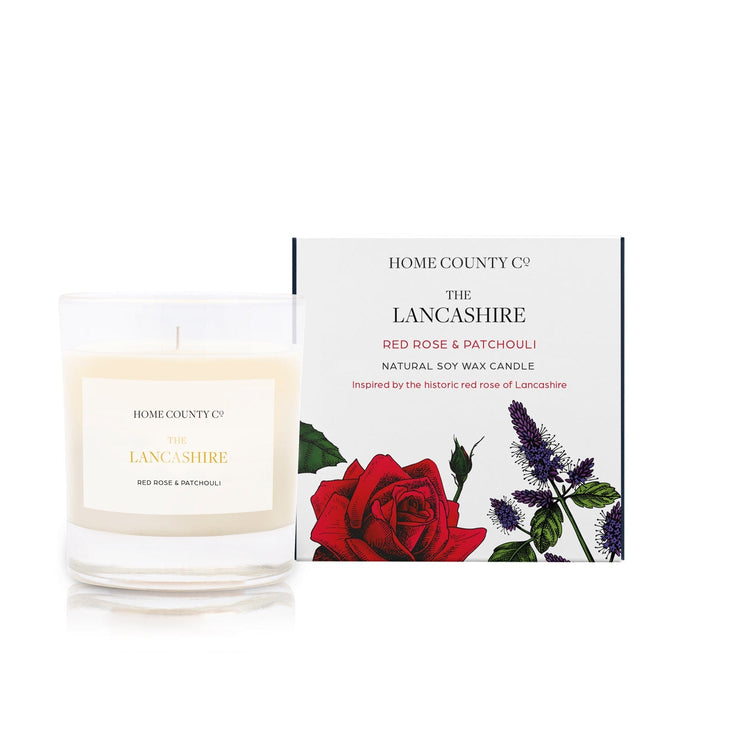 A red rose scented candle from the Home County Co is shown alight next to a red rose and its illustrated candle packaging
