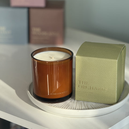 The Orchards candle from the Home County Co. is shown in its amber candle glass vessel next to the luxury gold foiled green box with apple emblem