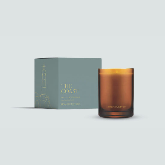 The Coast candle from the Home County Co. is shown in its amber candle glass vessel next to the luxury gold foiled blue box with lighthouse emblem
